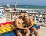 A happy couple enjoys a sunny day at the Sitges gay beach, sitting on a rainbow bench with the ocean in the background.