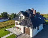 Nowoczesny dom z czarnym dachem, odpornym na warunki atmosferyczne, klasa korozyjności C4. Otoczony zielenią i zadbanym podjazdem.