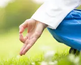 En hand i mudra, klädd i vitt och blått, vilar på gräset. Kirtan kriya forskning visar hur denna meditation kan påverka välbefinnandet.