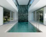 Piscina interior con pared de mármol verde veteado, rodeada de ventanales que dan a un jardín.