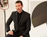 A man in a sharp black suit and polo shirt, a classic funeral outfit for men, poses with a thoughtful expression.