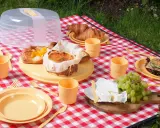 Co zabrać na piknik? Na czerwono-białej kratce leżą ciasta, croissanty, ser, winogrona i kanapka. Są też żółte talerze, kubeczki i sztućce.