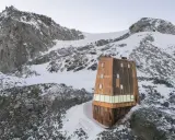 Il rifugio Sasso Nero, un'architettura moderna in legno, si erge solitario tra rocce e neve, dominato da imponenti vette montuose.