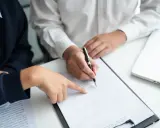 Dwie osoby przy biurku analizują dokument. Jedna wskazuje palcem na tekst, druga podpisuje umowę zlecenia.