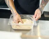 Ręce z tatuażami zagniatają ciasto w plastikowym pojemniku. To pierwszy krok, by dowiedzieć się, jak mrozić ciasto.