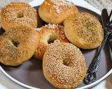 Sechs selbstgemachte Bagels auf einem Teller: zwei mit Sesam, zwei mit Kräutern und zwei mit grobem Salz bestreut. Ein Messer und eine Gabel liegen daneben.