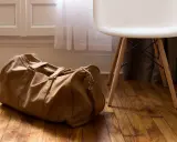 Brązowa torba podróżna leży na drewnianej podłodze obok białego krzesła. Idealna, by spakować wszystko, co potrzebne, gdy zastanawiasz się, co zabrać na żagle.