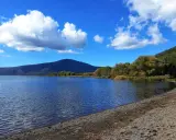 Spiaggia ghiaiosa e acque calme del lago di Vico, con montagne verdi e cielo azzurro punteggiato di nuvole bianche.