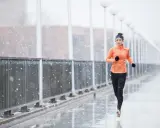 Donna corre sotto la pioggia su un ponte, con fiocchi di neve che cadono. Indossa abbigliamento sportivo arancione e nero.