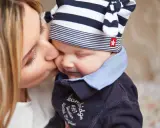 Delikatny pocałunek mamy, by obudzić dziecko. Uśmiechnięta twarz malucha w czapce w paski.
