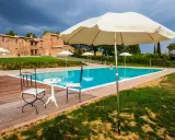 Piscina con vista su un casale in mattoni, perfetta per le tue vacanze in Toscana. Tavolino con sedie e ombrelloni pronti per rilassarsi.