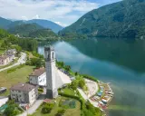 Torre campanaria affacciata sul lago di Corlo, con montagne verdi sullo sfondo e un villaggio pittoresco.