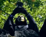 En figur med gyllene mask och svart kappa håller upp en bägare mot lövverket. Kanske är det "svarta solen geografens testamente" som avslöjas.