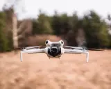Eine weiße Drohne A3, Modell Mini 4 Pro, schwebt über trockenem Gras. Im Hintergrund verschwimmen Bäume und eine bewölkte Himmel.