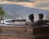 Coppia abbracciata in una vasca idromassaggio di legno, con lanterne accese e vista sulle montagne. Un momento di puro relax in Trentino.