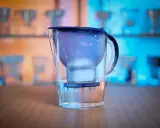 En vattenrenare för dricksvatten, en Brita-kanna, står på ett träbord. Bakgrunden visar fler liknande kannor i blått och orange ljus.