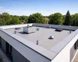 Nowoczesny dach płaski z elementami wentylacyjnymi i świetlikami. Widoczna jest estetyczna wysokość attyki, otoczona zielenią.