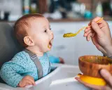 Ein Baby sitzt im Hochstuhl und bekommt von einer Hand mit einem Löffel Babybrei mit Fleisch gefüttert.