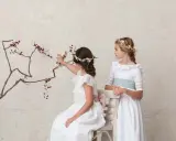 Dos niñas con vestidos blancos y coronas de flores, una sentada tocando una rama, la otra de pie. Peinados de trenza para niña de comunión.