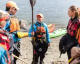 Grupp samlad på stranden, redo för paddling. En instruktör visar paddelteknik. Kajaker ligger i bakgrunden.