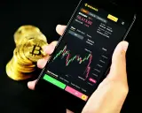 Ręka trzyma smartfon z wykresem kursu Bitcoina na platformie Binance. Obok stos złotych monet. Czy Binance czy Zonda? Decyzja należy do Ciebie.