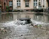 Woda z kanalizacji wydostaje się przez studzienkę, tworząc wodospad. Widać śmieci unoszące się na powierzchni.