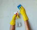 Ręce w żółtych rękawiczkach czyszczą ścianę. Jedna ręka trzyma butelkę z płynem, druga gąbkę. To nie jest grzyb w mieszkaniu, to czyszczenie.