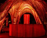 Struttura in bambù illuminata di rosso, con un bancone e un'installazione luminosa centrale. Atmosfera da house techno.