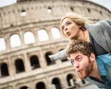 Coppia in una vacanza romantica a Roma, con il Colosseo sullo sfondo. Lui porta lei sulle spalle, entrambi con espressioni sorprese.