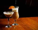 Dwa drinki z likierem kawowym i mlekiem: jeden w kieliszku martini, drugi w wysokiej szklance z pomarańczą.
