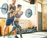 Frau hebt mit Trainer eine Langhantel. Sie konzentriert sich auf ihr exzentrik training, um Kraft aufzubauen.