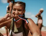 Läuferin beißt stolz in ihre Goldmedaille nach einem 10k Lauf. Andere Athletinnen im Hintergrund.