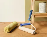 Färgburk och roller på trägolv, redo att måla på tapet.