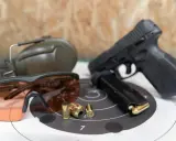 Pistolet, magazynek, łuski, okulary ochronne i słuchawki – kompletne akcesoria strzeleckie gotowe do użycia.