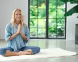 Kvinna i blå kläder sitter i lotusställning på en matta, med händerna i bön. Hon utstrålar lugn, redo för en stunds shaking meditation.