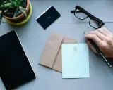 Ręka pisze ołókiem na jasnoniebieskiej papeterii, przygotowując list do Anglii. Obok zeszyt, okulary i koperta.