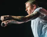 Macklemore na koncercie w Polsce, z mikrofonem w dłoni, w białej koszuli i z tatuażami na ramionach.