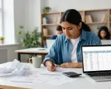 Uczennica pochylona nad zeszytem, obok kalkulator i laptop z tabelą wyników. Intensywna nauka do egzaminów **international baccalaureate**.
