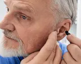 Starszy mężczyzna z siwą brodą próbuje włożyć czarny zatyczkę do ucha, kiedy aparat słuchowy nie pomaga.