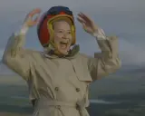 Glad Trine Dyrholm i pilotmössa och trenchcoat vinkar glatt. En scen från filmer med Trine Dyrholm.
