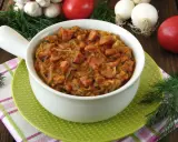 Pyszny bigos z młodej kapusty z kiełbasą, podany w białej misce na zielonym talerzyku. W tle pomidory, czosnek i koperek.
