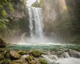 Cascata maestosa nei Laghi di Plitvice, con acqua turchese e rocce muschiose. La luce solare filtra tra gli alberi, creando un'atmosfera magica.