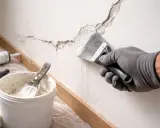 Ręka w rękawicy nakłada gładź na pęknięcia na ścianie. W pobliżu wiaderko z masą szpachlową i narzędzia. Dopuszczalne rysy na tynku zostaną naprawione.