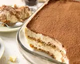 Pyszne tiramisu bez jajek w szklanej formie, posypane kakao. Obok porcja deseru na talerzyku.