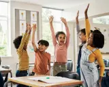Fröhliche Kinder mit erhobenen Armen in einem Klassenzimmer. Sie nehmen begeistert an Kennenlernspielen für die Grundschule teil.