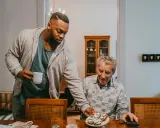 Un jeune homme sert une tasse à un aîné, partageant un moment de **colocation intergénérationnelle** autour d'un plateau de tartines.