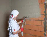 Pracownik w białym ubraniu nakłada tynk agregatem na ścianę z cegły. Obliczanie, ile wiader tynku na 200m2, to klucz do efektywnej pracy.