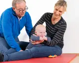 Ein Mann und eine Frau üben mit einem Baby auf einer roten Matte. Das Baby greift nach seinen Fingern. kg zns kinder.