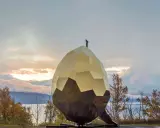 Solägg i Artipelag, en gyllene, fasetterad skulptur som liknar ett ägg, står mot en dramatisk himmel vid en sjö.