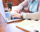 Osoba pisząca na laptopie, z zegarkiem na ręce, obok notatnika i kubka z herbatą.
